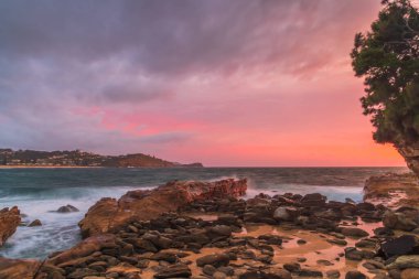 Rüzgârlı gündoğumu deniz manzarası bulutlarla kaplı gökyüzü, kayalar ve dalgalar Orta Sahildeki Avoca Sahili, NSW, Avustralya.