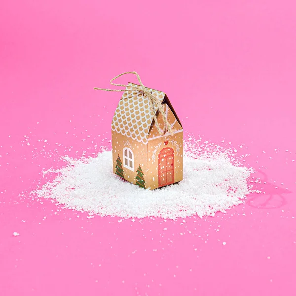 Karda şirin bir ev, pastel pembe arka plan. Gerçeküstü kış sezonu yaratıcı konsepti. Noel veya Yeni Yıl atmosfer afişi veya kart tasarımı.