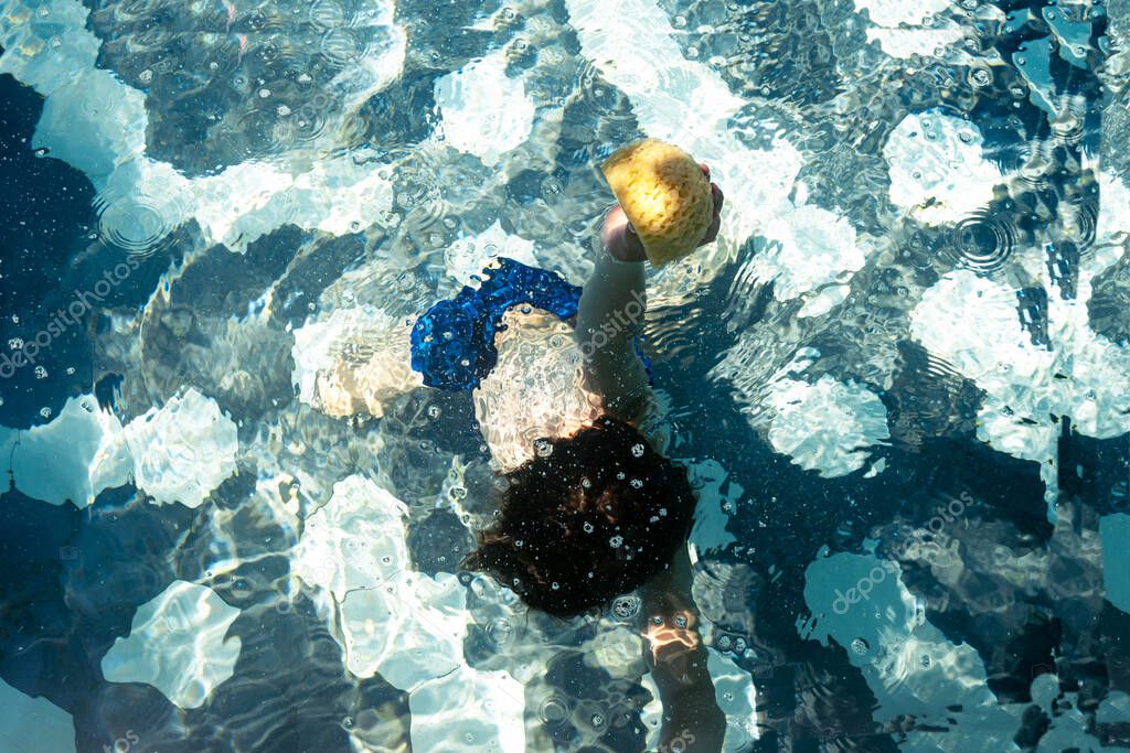 A young boy diving and taking out of the swimming pool a sea sponge. Creative concept for hotel resort or travel agency advertisement. Design for summer holidays banner