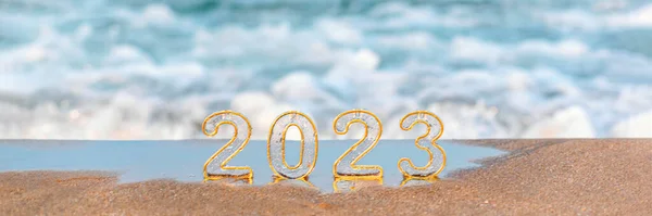Happy new year 2023 wide banner, background. Numbers 2023 on golden sand of beach and blurry sea wave. New Year's Eve holidays celebration, travel, vision, dreams, goals concept, copy space
