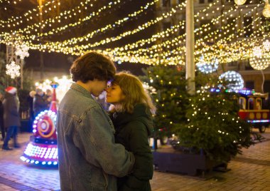 Kış tatili, birbirine aşık mutlu genç çift akşamları Noel pazarında buluşuyor. Arka planda bir sürü Noel ışığı var. Yumuşak odaklanma, gürültü. Tatil atmosferi