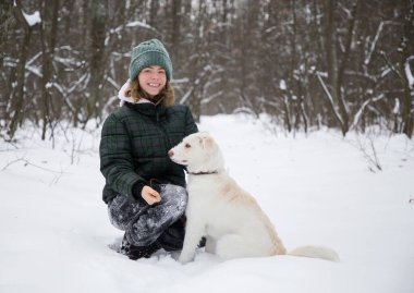 16-17 yaşlarındaki tatlı gülümseyen genç kız beyaz köpeğiyle karlı ormanda yürüyor. Soğuk kış mevsimi. Aktif yaşam tarzı, sevgili evcil hayvanla yürümek, her gün neşe
