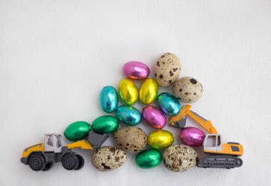 models of toy excavator and loader, lot of chocolate eggs in colorful wrappers, quail eggs on light background. concept of business congratulations on Easter holiday for construction companies.