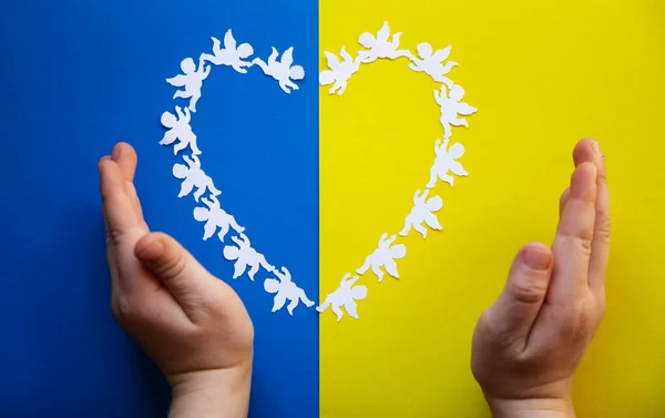Heart, laid out of paper angels on yellow blue background in colors of Ukrainian flag, children\'s hands. children of Ukraine are against war, Ukrainians want peace. concept of support, faith, hope