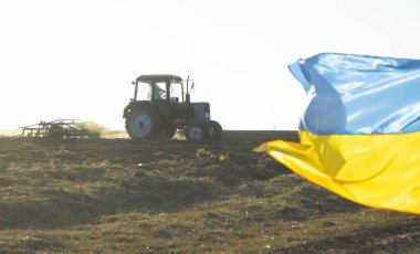 Tarlayı eken traktör, tarım arazisi. Sarı-mavi Ukrayna bayrağı yan yana dalgalanıyor. İçinde metin olan bir resim. Savaş sırasında Ukrayna 'da mevsimsel tarım işleri. Ukrayna 'yı Destekle