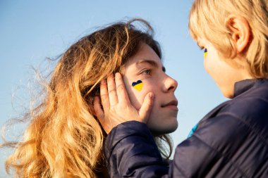 Kadının yüzü küçük bir çocuğun eliyle tutulur, Ukrayna bayrağının kalbi yanağına çizilir. Aile, birlik, teselli. Savaş boyunca birbirinize iyi bakın ve destek olun.