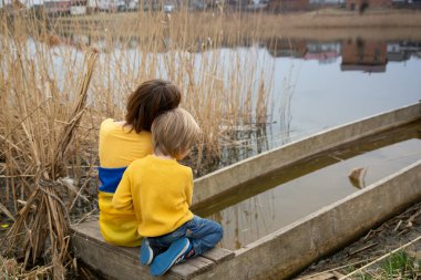Küçük bir çocuk ve bir kadın, sırtları Ukrayna bayrağı taşıyan mavi-sarı elbiseli bir gölün kıyısında oturuyorlar. Aile, mülteciler, birlik, destek, vatanseverlik, nostalji. Ukrayna 'nın yanında dur