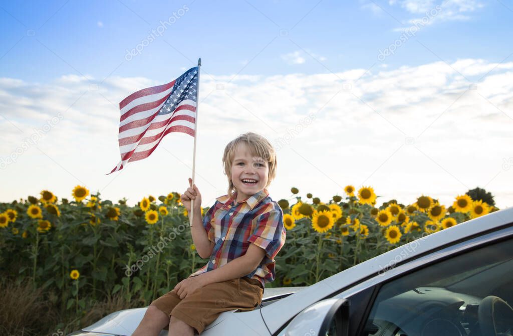 smiling boy of 5 years old sits on white car with American flag in his hand against backdrop of blooming field of sunflowers. Independence Day of United States of America. Pride, Patriotism, Freedom