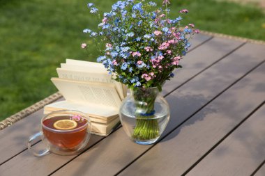 Masada bir fincan çay, açık bir kitap ve bahçede cam vazoda çiçeklerle dolu bir hayat. Güzel bir sabah fotoğrafı. Dinlenme zamanı. Zevkle kitap okuyorum. İyi bahar günleri.