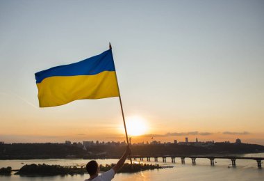 Büyük Ukrayna bayrağı günbatımı gökyüzü ve şehir manzarasının zemininde dalgalanıyor. Kyiv 'de bir adam evin çatısında duruyor ve bayrak taşıyor. Ulusal özgürlük ve bağımsızlık sembolü. Savaşı durdurun.