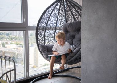 6 yaşında bir çocuk, büyük pencerelerin yakınındaki sallanan bir sandalyede oturmuş kitap okuyor. Dijital detoks konsepti, evde eğitim