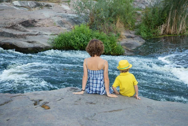 Two unrecognizable people - woman and child sit with their backs against backdrop of beautiful landscape and flow of water from mountain river. pleasant family pastime for weekend. travels. tourism