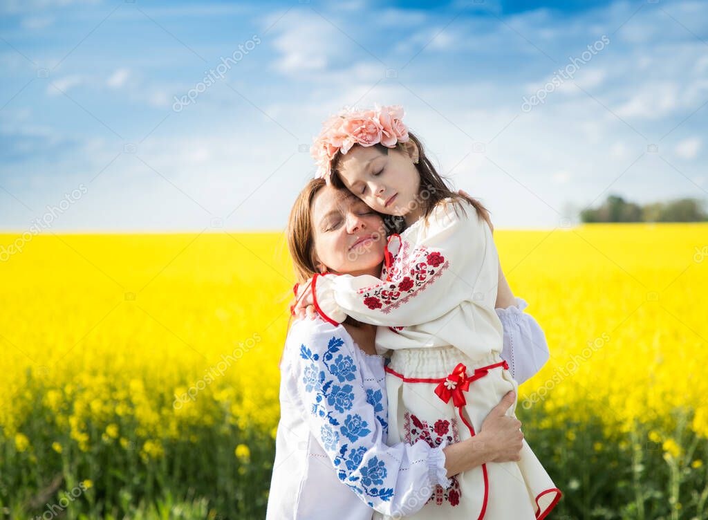 Ukrainian family, mother and daughter, in embroidered shirts, gently hug among blooming yellow rapeseed field. unity, security. independence Day. Ukrainians ask for support and peace for Ukraine.
