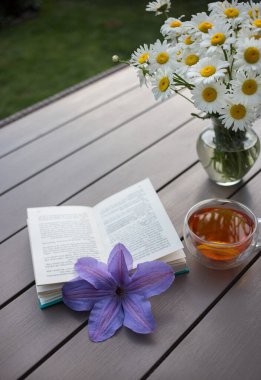 Masada bir fincan çay, açık bir kitap, mavi klematis çiçeği ve vazoda bir buket papatya. Sakin bir sabah fotoğrafı. Dinlenmek ve çay içmek. Zevkle kitap oku. Sıcak, sade bir ortam.