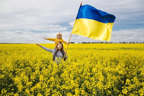 黄色の菜の花畑の中でウクライナの大きなサテンの旗を持つ肩の上に彼女の息子と母親 ウクライナへの栄光 愛国心の教育です 国家独立 戦争を止めて — ストック写真
