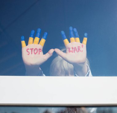 Çocuğun elleri cam camında, sarı-mavi renkte boyanmış, kırmızı STOP SAVAŞ yazılı. Rusya 'nın Ukrayna' yı işgali, çocukların savaşa karşı olması. Ukrayna 'dan uzak dur. Ukraynalılara yardım et