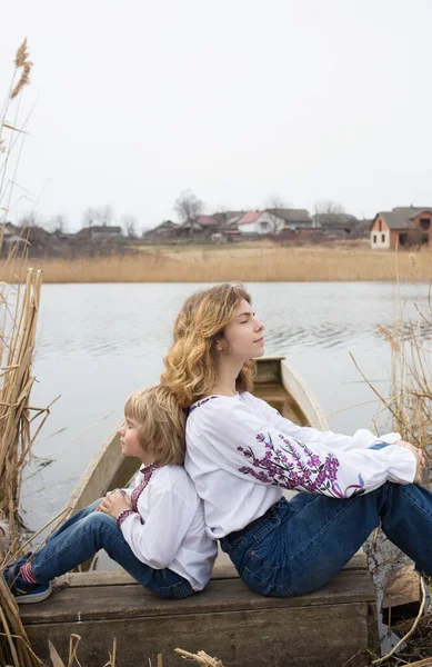 little boy and a young beautiful woman in embroidered national clothes are sitting back to back on the shore of the lake. Family, refugees, unity, support, sadness. Ukrainians are against the war