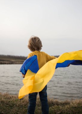 unrecognizable boy stands with his back on the shore of a lake with a fluttering blue and yellow Ukrainian flag. pride to be Ukrainian. People are against war. stand with Ukraine. Refugees, nostalgia