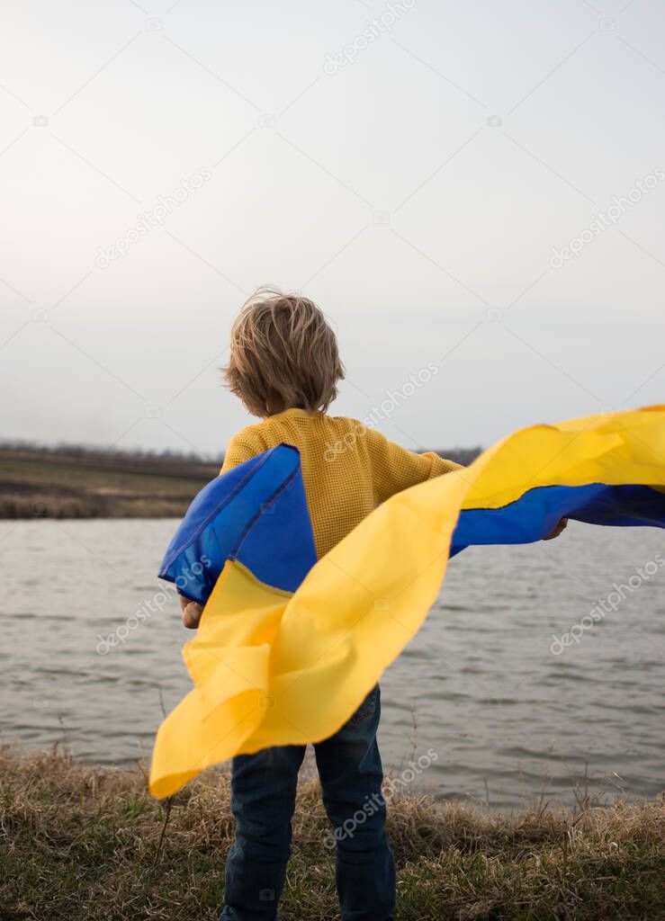 unrecognizable boy stands with his back on the shore of a lake with a fluttering blue and yellow Ukrainian flag. pride to be Ukrainian. People are against war. stand with Ukraine. Refugees, nostalgia