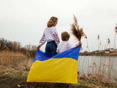 little boy and young woman stand with their backs on shore of lake with blue yellow Ukrainian flag behind them. Family, refugees, unity, solidarity, support, patriotism. Ukrainians are against war