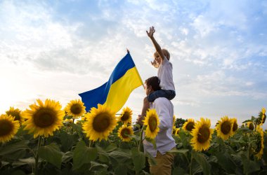 dad with son, who sits on his shoulders, with large flag of Ukraine among a blooming field with sunflowers. Patriotic education. Pride, faith in victory. Independence Day. Ukrainians against war
