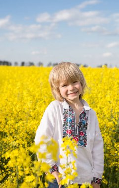cute smiling Ukrainian boy of 6 years old in a traditional embroidered shirt stands among a yellow flowering rapeseed field. Children for Peace. Support Ukraine. Proud to be Ukrainian. Sunny day