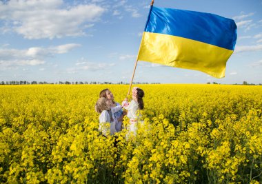 ukrainian family mother, son and daughter in embroidered shirts stand among blooming yellow rapeseed field with large flag of Ukraine. Sunny day. unity support. independence Day. Children against war.