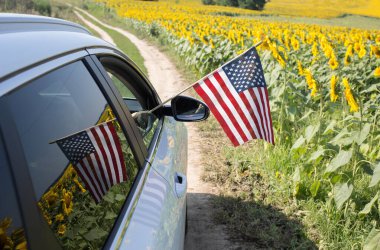 American flag in hand is stuck out of car window. Reflection of flag and blooming field of sunflowers in window. vacation, travel. Independence Day of United States of America. Pride, Patriotism