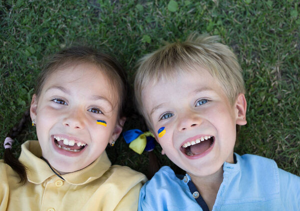 Лица двух смеющихся счастливых детей, мальчик и девочка 6-7 лет в желтых и синих футболках, лежат на траве. Вид сверху. День независимости. Дети Украины за мир. Поддержка Украины