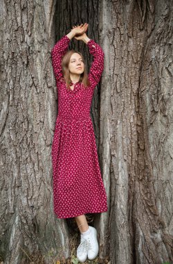 Full length portrait of a girl in a long burgundy dress posing in the shade of an oak tree trunk. Loneliness, contemplation, solitude, fantasies alone