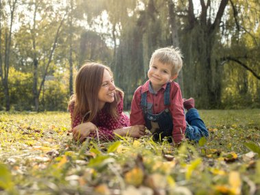 Smiling woman and little boy lie on grass on a sunny day strewn with autumn leaves. Mother and son are relaxing in the park at the weekend. Family outdoor recreation, pastime, happy motherhood