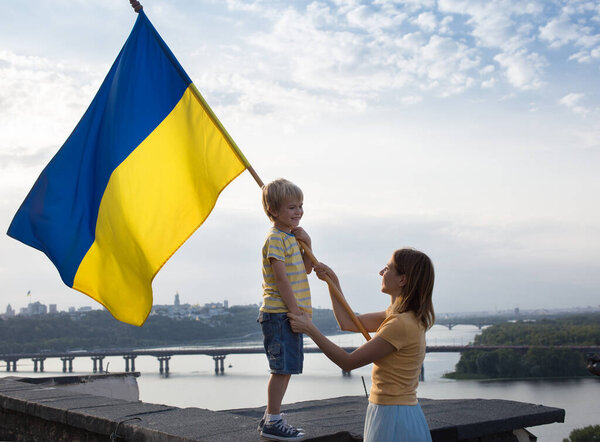 Mom and little son stand high on the roof of the house against the background of the sky and the Dnieper River in Kyiv. Patriotism, drawing attention to the war in Ukraine. Support Ukraine