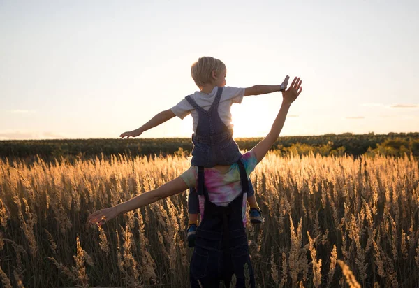 unrecognizable child sits on shoulders of mom and walks along meadow illuminated by sunset light. Happy childhood, have fun together on walk. Forward to bright future. back view. Backlight soft focus