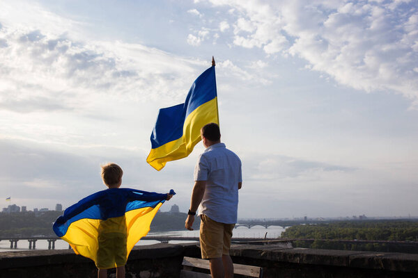 мужчина и ребенок стоят спиной на высокой крыше дома в Киеве над Днепром с двумя флагами Украины против неба. Патриотическое воспитание. Гордость, свобода. Поддержка Украины
