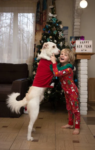 クリスマスの赤いパジャマの子供と白い犬はクリスマスツリーの前の家の部屋で抱き合っています 幸せな家族の冬の休暇のコンセプト 居心地の良いお祝い陽気な雰囲気 — ストック写真