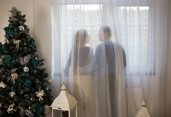 Silhouettes of couple in love, man woman, standing with backs near window behind curtain. decorated Christmas tree in room. atmosphere of holiday, love, tenderness, comfort. Theme Christmas morning.