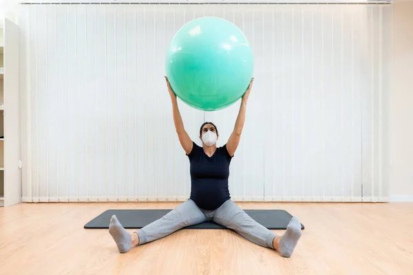 Close up of a pregnant woman with face mask sitting on floor doing pilates exercises with a ball — Stock Photo, Image