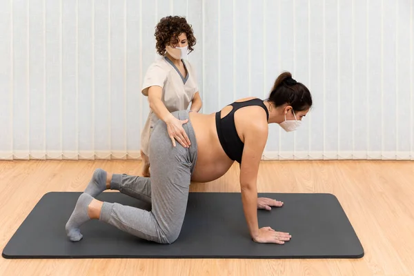 Yüzü maskeli hamile bir kadın mindere diz çöküp fizyoterapistin yardımıyla yoga yapıyor. — Stok fotoğraf