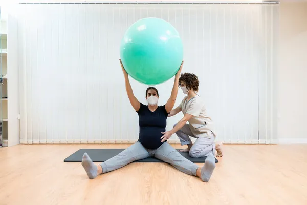 Yüzü maskeli hamile bir kadın, fizyoterapistinin yardımıyla yerde pilates egzersizleri yapıyor. — Stok fotoğraf