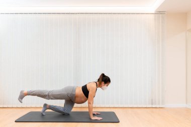 Yüzü maskeli hamile bir kadın paspasın üzerine çömelmiş evde fotokopi alanı olan bir yerde pilates yapıyor.
