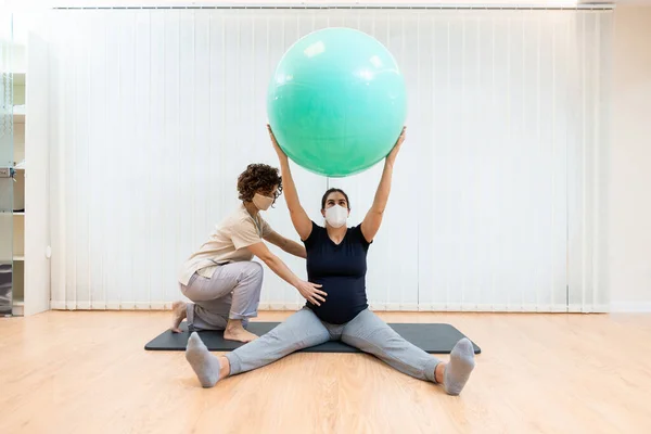 Yüzü maskeli hamile bir kadın, fizyoterapistin yardımıyla pilates egzersizleri yapıyor. — Stok fotoğraf