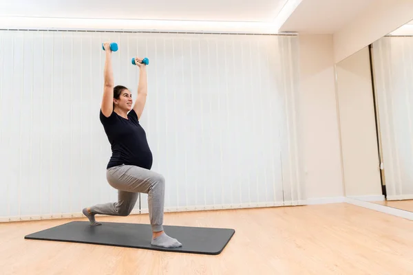 Neşeli bir hamile, hafif dambıllarla hamleler yapıyor sağlık merkezindeki bir paspasın üzerinde egzersiz yapıyor. — Stok fotoğraf