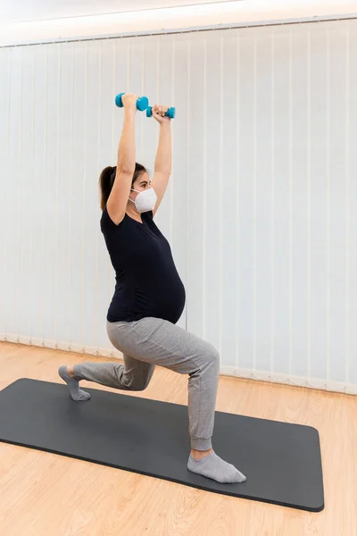 A pregnant with face mask doing lunges with light dumbells exercises on a mat at home