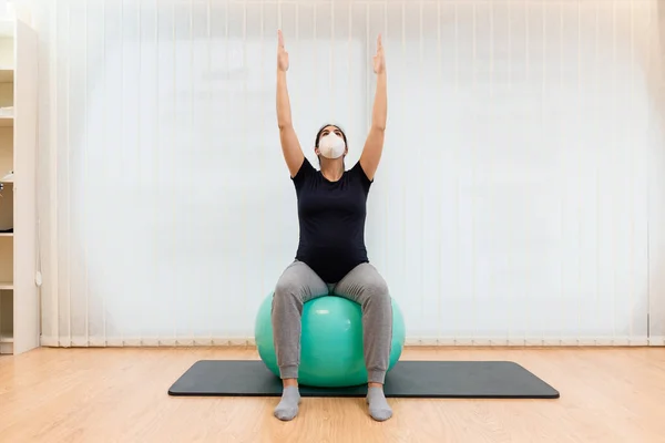Yüz maskesi takan hamile bir kadın pilates egzersizleri yapıyor spor topunun üzerinde oturuyor ve kolları yukarı doğru gerilmiş. — Stok fotoğraf