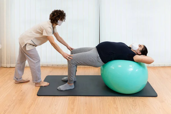 Una mujer embarazada acostada boca arriba en una pelota de fitness haciendo ejercicios con la ayuda de su fisioterapeuta —  Fotos de Stock