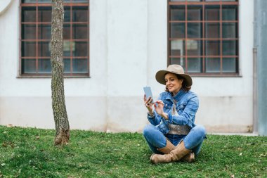 Dışarıdaki yeşil çimlerin üzerinde rahatça otururken cep telefonu kullanan bir kadın..