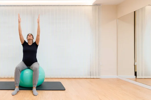 Hamile bir kadın pilates egzersizi yapıyor spor topunun üzerinde oturuyor, kolları yukarı doğru gerilmiş ve yukarı bakıyor. — Stok fotoğraf
