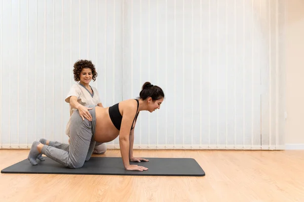 A pregnant woman kneeling on a mat doing yoga with help of physiotherapist in spa center — Stok fotoğraf