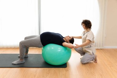 Yüzü maskeli hamile bir kadının, yerde oturup fizyoterapistle birlikte pilates egzersizleri yapmasını.