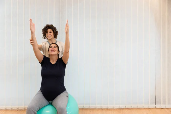 Hamile bir kadın fizyoterapistin yardımıyla bir fitness topunun üzerinde oturuyor. — Stok fotoğraf